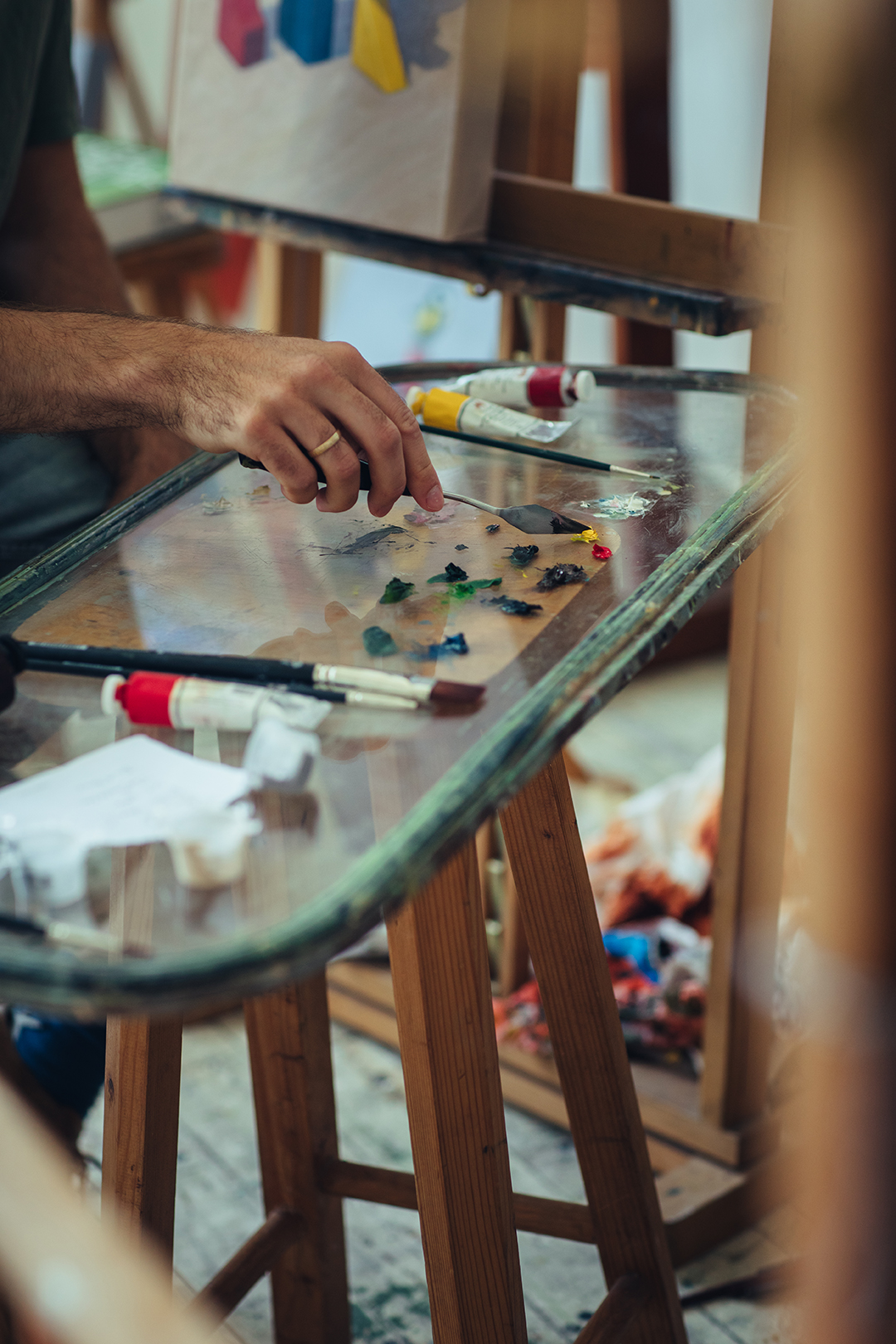 Een close-up van Rafaels hand boven een glazen schilderspalet vol verfkleuren. Op de werktafel liggen verfspullen en penselen tussen de sporen van eerdere schilderexperimenten.